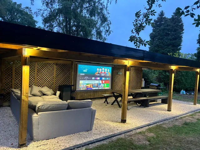 Covered timber garden room at dusk, with lights, a sofa and a television